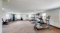 Located at 6533 Cedar Furnace Circle in Glen Burnie, Maryland, this single-floor fitness center offers a comprehensive workout facility designed to accommodate a wide range of exercise needs. The main cardio and strength training area is generously sized, featuring multiple treadmills, elliptical machines, and stationary and spin bikes, along with a multi-function cable weight machine and a workout bench. Wall-mounted flat-screen televisions provide entertainment during workouts, while recessed lighting and multiple windows allow natural light to fill the space. The patterned carpet flooring adds a comfortable feel underfoot throughout the cardio zone. A dedicated studio room offers a bright, open environment with light wood-style flooring, floor-to-ceiling mirrors, purple accent walls, and a modular storage unit—ideal for yoga, stretching, or group fitness classes. The studio benefits from natural light through blinded windows and features a clean, motivating atmosphere. A conveniently located restroom with tile flooring, a wall-mounted sink, and a large mirror rounds out the facility. A water cooler station and clearly marked emergency exits reflect the center's attention to member comfort and safety throughout.