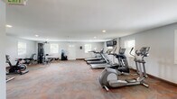 Located at 6533 Cedar Furnace Circle in Glen Burnie, Maryland, this single-floor fitness center offers a comprehensive workout facility designed to accommodate a wide range of exercise needs. The main cardio and strength training area is generously sized, featuring multiple treadmills, elliptical machines, and stationary and spin bikes, along with a multi-function cable weight machine and a workout bench. Wall-mounted flat-screen televisions provide entertainment during workouts, while recessed lighting and multiple windows allow natural light to fill the space. The patterned carpet flooring adds a comfortable feel underfoot throughout the cardio zone. A dedicated studio room offers a bright, open environment with light wood-style flooring, floor-to-ceiling mirrors, purple accent walls, and a modular storage unit—ideal for yoga, stretching, or group fitness classes. The studio benefits from natural light through blinded windows and features a clean, motivating atmosphere. A conveniently located restroom with tile flooring, a wall-mounted sink, and a large mirror rounds out the facility. A water cooler station and clearly marked emergency exits reflect the center's attention to member comfort and safety throughout.