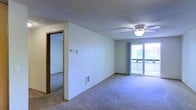 This single-floor residence offers a straightforward and functional layout designed for comfortable everyday living. The main living area is generously sized, finished with wall-to-wall carpeting that adds warmth underfoot. A ceiling fan is centrally positioned overhead, providing both air circulation and ambient lighting. A textured ceiling runs throughout the space, adding a classic residential character.

Natural light fills the living area through a large sliding glass door, which opens to a private balcony with views of the surrounding greenery and neighboring buildings — making it a pleasant spot for outdoor relaxation. Sheer curtains frame the glass door, allowing light to filter through softly.

The neutral, light-toned walls provide a clean and versatile backdrop throughout the space. A doorway visible from the living area leads to an adjacent room, suggesting a connected layout that facilitates easy movement between spaces. The unit also features built-in wall fixtures, including a thermostat and additional utility access points, indicating practical infrastructure throughout.

Overall, the space presents a clean, move-in-ready environment with a calm, neutral aesthetic, well-suited for those seeking a functional and comfortable single-level living arrangement with the added benefit of outdoor access.