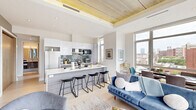 This contemporary 2-bedroom, 3-bathroom residence showcases a thoughtfully designed open-concept layout that maximizes both space and natural light. The combined kitchen and living area serves as the heart of the home, anchored by dramatic floor-to-ceiling windows that frame sweeping urban views and flood the space with daylight. Exposed concrete ceilings with warm wood beam accents lend an industrial-chic character throughout.

The kitchen is outfitted with sleek two-tone cabinetry — pairing high-gloss white uppers with wood-grain lowers — a subway tile backsplash, stainless steel appliances, and a generous island with bar seating, ideal for casual dining and entertaining. Light hardwood flooring runs throughout the main living areas, complementing the bright, neutral palette.

The primary bedroom offers a tranquil retreat with an open closet, city views through large windows, and access to a spa-inspired en suite bathroom featuring a glass-enclosed walk-in shower adorned with chevron-patterned tile. The second bedroom is equally comfortable, with exposed concrete ceilings and ample natural light. Additional highlights include a hallway with built-in coat hooks, a dedicated laundry area, two closets, and a private balcony perfect for enjoying the urban skyline.