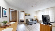 Located along the Katy Freeway in Houston, Texas, this well-appointed two-bedroom, two-bathroom suite offers a functional and comfortable single-floor layout. Light wood-look flooring runs throughout the space, creating a cohesive and clean aesthetic.

The open-concept living area is anchored by a gray sofa and features sheer curtained windows that allow ample natural light to fill the room. A peninsula counter with a granite-style surface separates the living space from the kitchen, which is equipped with warm wood cabinetry, granite countertops, a full-size refrigerator, electric range, microwave, and a sink — providing everything needed for self-sufficient living.

The primary bedroom includes a wall-mounted television, a dedicated workspace with a desk and chair, and access to natural light through a window with sheer curtains. A second bedroom also features a wall-mounted TV and a large bed with clean, neutral bedding. Both bedrooms are accessed via classic six-panel wood doors.

The two bathrooms are neatly designed: one features an open-shelf vanity with a rectangular undermount sink, a wood-framed mirror, and stone-look countertops, while the other includes a tub/shower combination with marble-tile surround. A dedicated closet with a full hanging rod provides additional storage throughout the suite.