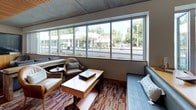 This single-floor lounge space showcases a thoughtfully designed interior that balances contemporary architecture with comfortable, functional furnishings. A standout feature is the striking slatted wood ceiling, which adds warmth and acoustic character to the space, contrasting elegantly with the exposed concrete elements throughout.

A continuous bank of floor-to-ceiling windows runs along one side of the room, flooding the interior with natural light and offering open views of the surrounding streetscape and greenery. Multiple seating arrangements are distributed throughout, including plush gray sofas, leather club chairs, and built-in banquette-style seating, each grouping centered around low wooden coffee tables atop a vibrant, multi-toned patterned carpet.

Entertainment is well-provided for with a large wall-mounted flat-screen television accompanied by warm wood cabinetry nearby. A full-length shuffleboard table anchors one end of the lounge, adding a recreational element to the space.

The kitchenette area is equipped with warm maple cabinetry, granite-style countertops, a deep utility sink, an espresso/coffee machine, and an ice maker — all finished beneath the same elegant slatted wood ceiling. The polished concrete flooring in this zone complements the modern industrial aesthetic while remaining practical and easy to maintain.