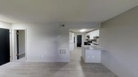 This single-floor residence offers a clean, modern aesthetic defined by an open-concept floor plan that seamlessly connects the living area to the kitchen. Light gray walls and a textured ceiling complement the smooth, light-toned hardwood-style flooring that runs throughout, creating a cohesive and airy feel.

The kitchen is a standout feature, outfitted with crisp white cabinetry that extends to the upper walls, providing ample storage space. A peninsula with a granite-style countertop serves as a natural divider between the kitchen and living area, offering additional prep space and the opportunity for casual seating. Black appliances, including a refrigerator and range, add a bold contrast against the white cabinetry, while a stainless steel sink with an overhead window brings in a touch of natural light. Under-cabinet range ventilation and a flush-mount ceiling light ensure the kitchen is both functional and well-lit.

The living area is open and versatile, featuring a centrally located HVAC vent and a thermostat panel on the wall, ensuring year-round climate comfort. A secondary floor-level air return vent is also present, reflecting a well-integrated HVAC system. The overall ambiance is bright, neutral, and move-in ready, ideal for a variety of interior styles.