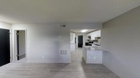 This single-floor residence offers a clean, modern aesthetic defined by an open-concept floor plan that seamlessly connects the living area to the kitchen. Light gray walls and a textured ceiling complement the smooth, light-toned hardwood-style flooring that runs throughout, creating a cohesive and airy feel.

The kitchen is a standout feature, outfitted with crisp white cabinetry that extends to the upper walls, providing ample storage space. A peninsula with a granite-style countertop serves as a natural divider between the kitchen and living area, offering additional prep space and the opportunity for casual seating. Black appliances, including a refrigerator and range, add a bold contrast against the white cabinetry, while a stainless steel sink with an overhead window brings in a touch of natural light. Under-cabinet range ventilation and a flush-mount ceiling light ensure the kitchen is both functional and well-lit.

The living area is open and versatile, featuring a centrally located HVAC vent and a thermostat panel on the wall, ensuring year-round climate comfort. A secondary floor-level air return vent is also present, reflecting a well-integrated HVAC system. The overall ambiance is bright, neutral, and move-in ready, ideal for a variety of interior styles.