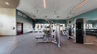 Located at 1715 Lake Michigan Dr in Valparaiso, Indiana, this fitness center offers a generously sized, single-floor exercise facility designed to support a comprehensive workout experience. The space is thoughtfully divided into distinct zones, each tailored to different training needs.

The cardio area features a row of treadmills, elliptical trainers, and stationary bikes positioned along a wall of large windows, allowing abundant natural light to fill the room and providing views of the surrounding exterior. Roller shades offer adjustable light control, while ceiling fans and modern linear pendant lighting enhance comfort and visibility throughout.

The strength training zone is equipped with a full rack of dumbbells, weight benches, cable machines, and multi-use strength stations, all set against floor-to-ceiling mirrors that visually expand the space. Rubberized flooring in this area provides traction and durability.

A dedicated functional fitness area features open floor space with carpet and hardwood-style flooring, along with stability balls, medicine balls, resistance equipment, and a squat rack. Soaring ceilings with an arched detail amplify the sense of openness in this zone.

A separate cycling and spin area includes stationary bikes positioned beneath tall windows with a wall-mounted television. The space's teal accent walls, warm wood details, and clean architectural lines create a modern, energizing atmosphere ideal for fitness and wellness.