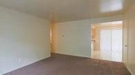 This single-floor space presents a straightforward, functional layout designed for comfortable everyday living. The main living area is finished with wall-to-wall carpeting in a neutral tone, providing a warm underfoot feel, while plain white walls and a smooth ceiling create a clean, understated backdrop. An open doorway connects the living area directly to the kitchen, which features light wood cabinetry, a countertop bar area, and tile flooring that contrasts with the carpeted living space. Natural light filters into the kitchen through vertical blinds covering what appears to be a sliding glass door or large window, brightening that section of the unit. A secondary doorway is also visible from the living area, suggesting access to additional rooms or a hallway beyond. The overall color palette is soft and neutral, with beige walls and light wood accents contributing to a cohesive, muted aesthetic. The space is modest in scale but benefits from an open sightline between the main living and kitchen areas, lending a sense of connectivity throughout the floor plan.