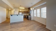 This single-floor residence is designed with a contemporary aesthetic, combining functional layout with modern finishes. The open-concept living and dining area features wood-look hard-surface flooring, neutral gray walls, and an exterior door that allows natural light to filter in through slatted blinds. A wall-mounted HVAC unit and ceiling vents ensure year-round climate control throughout the main living space.

The kitchen is well-appointed with dark wood-toned cabinetry, stainless steel appliances including a gas range, microwave, and refrigerator, and light-colored countertops. A peninsula with pendant lighting above serves as a natural divider between the kitchen and dining area, which is further brightened by a large bank of windows fitted with white blinds.

The bedrooms are finished with plush, neutral-toned carpeting and feature clerestory-style windows that introduce natural light while maintaining privacy. Each room includes ample wall space and ceiling vents for ventilation.

Two full bathrooms mirror each other in design, each offering dark cabinetry vanities with white countertops, wood-look tile flooring, large mirrors, and a shower-tub combo with a curved curtain rod. Both bathrooms maintain a clean, cohesive aesthetic consistent with the rest of the unit.
