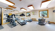 Located at 570 Stowell Drive in Rochester, NY, this fitness center offers a comprehensive workout facility spread across a single floor. The space is thoughtfully organized into distinct exercise zones, each designed to support a full range of fitness activities.

The main cardio area is generously sized and features multiple treadmills, an elliptical trainer, and a stationary bike, all positioned on protective floor mats. Full-length mirrors line the walls, creating an open and spacious feel while allowing users to monitor their form. Warm wood-trimmed ceiling light panels provide bright, even illumination throughout, and several windows with wood-framed surrounds bring in natural light and views of the surrounding greenery.

A dedicated strength training room houses a multi-station cable machine, a flat weight bench, and a tiered dumbbell rack stocked with a wide selection of free weights. A water cooler is conveniently located nearby. This area also features a skylight-style ceiling panel and mirrored walls that enhance the sense of space.

Additional amenities include a wall-mounted television, a bulletin board with posted fitness resources, a large floor fan, and wall-mounted hooks for personal items. The facility is Wi-Fi equipped, as indicated by posted signage. Neutral carpet flooring, clean white walls, and consistent warm-toned wood accents throughout give the space a cohesive, welcoming aesthetic suited for focused, effective workouts.