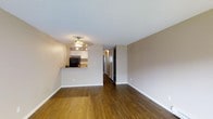 This 1-bedroom, 1-bathroom unit offers a well-organized single-floor layout designed for efficient and comfortable living. The open living area features warm hardwood flooring that extends throughout the main space, creating a cohesive and inviting atmosphere. Neutral-toned walls and a textured ceiling complement the understated aesthetic, while a ceiling fan with integrated lighting provides both comfort and illumination. The kitchen is positioned adjacent to the living area and is defined by a raised breakfast bar that serves as a natural divider between the two spaces. White cabinetry lines the kitchen walls, offering ample storage, while black appliances add a contrasting accent. A granite-style countertop completes the kitchen's functional design. A hallway leading from the living area connects to the bedroom and bathroom, with a sliding-door closet visible along the corridor, providing convenient storage access. The overall layout is compact yet thoughtfully arranged, making practical use of the available square footage. The neutral color palette and warm flooring tones give the space a welcoming character that suits a variety of décor styles.
