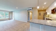 This single-floor residence offers a thoughtfully designed open-concept layout that seamlessly connects the kitchen and living areas. The kitchen is a standout feature, showcasing white shaker-style cabinetry extending to the ceiling, granite countertops, stainless steel appliances including a refrigerator, dishwasher, range, and microwave, as well as warm hardwood-style flooring and a peninsula with pendant lighting overhead. The spacious living area is carpeted and bathed in natural light from a large multi-pane window and sliding glass door that provides views of the surrounding greenery. A wall-mounted mini-split system provides efficient climate control throughout the main living space. The bedrooms are generously sized, each featuring plush carpeting, neutral tones, and windows that invite natural light. One bedroom also includes its own mini-split unit. The full bathroom is cleanly finished with a granite-topped white vanity, a tub-shower combination with a rainfall showerhead, and wood-look flooring. A dedicated in-unit laundry area with a stacked washer and dryer adds everyday convenience. Recessed lighting throughout enhances the bright, clean ambiance of this move-in-ready space.