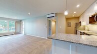 This single-floor residence offers a thoughtfully designed open-concept layout that seamlessly connects the kitchen and living areas. The kitchen is a standout feature, showcasing white shaker-style cabinetry extending to the ceiling, granite countertops, stainless steel appliances including a refrigerator, dishwasher, range, and microwave, as well as warm hardwood-style flooring and a peninsula with pendant lighting overhead. The spacious living area is carpeted and bathed in natural light from a large multi-pane window and sliding glass door that provides views of the surrounding greenery. A wall-mounted mini-split system provides efficient climate control throughout the main living space. The bedrooms are generously sized, each featuring plush carpeting, neutral tones, and windows that invite natural light. One bedroom also includes its own mini-split unit. The full bathroom is cleanly finished with a granite-topped white vanity, a tub-shower combination with a rainfall showerhead, and wood-look flooring. A dedicated in-unit laundry area with a stacked washer and dryer adds everyday convenience. Recessed lighting throughout enhances the bright, clean ambiance of this move-in-ready space.