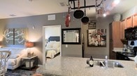 This single-floor residence offers a thoughtfully designed layout that blends functionality with a warm, inviting ambiance. The kitchen serves as a central hub of the space, showcasing striking granite countertops that provide ample prep and serving surface. A decorative ceiling-mounted pot rack adds a culinary flair while keeping cookware conveniently accessible, and the kitchen is equipped with a stainless steel sink to complete its practical setup.

The open sightlines from the kitchen connect directly to the bedroom, which features a classic upholstered headboard, neutral bedding, and warm lamp lighting that creates a relaxing retreat. Gray-toned walls flow consistently throughout the space, lending a cohesive and modern aesthetic. A framed mirror adds depth to the wall decor, complemented by expressive wall art that injects personality and character into the interior.

The layout promotes a sense of openness and connectivity between living areas, making the space feel welcoming and easy to navigate. Recessed ventilation and thoughtful lighting placement contribute to a comfortable indoor environment. Overall, this residence balances practical design elements with stylish touches, resulting in a space that feels both livable and visually engaging.