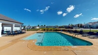 Located at Ashford Villa in Chattanooga, Tennessee, this single-level outdoor amenity space serves as a central gathering hub for the community. At its heart is a generously sized swimming pool with clearly marked depth indicators, accessible entry points including handrail-equipped steps, and crystal-clear blue water that reflects the open Tennessee sky.

The expansive concrete pool deck surrounds the pool on all sides, providing ample space for lounging. Numerous chaise lounge chairs and patio seating sets are arranged throughout the deck, offering both open-air sunbathing and shaded retreat options. A covered porch area with a pitched roof and white exterior columns extends from the adjacent clubhouse building, providing a comfortable shaded zone furnished with additional lounge seating.

The pool area is enclosed by a black wrought-iron fence with decorative stone landscaping at its base, offering both security and aesthetic appeal. Mature evergreen trees add vertical greenery to the setting, while well-maintained lawns and community residences are visible beyond the fence line, reinforcing the neighborhood atmosphere.

The overall ambiance is bright, open, and inviting, benefiting from abundant natural sunlight and wide open skies. This outdoor amenity space is thoughtfully designed to foster community enjoyment and relaxation in a welcoming residential setting.