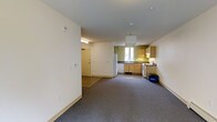 Located at 2 Gile Hill in Hanover, New Hampshire, this single-floor unit offers a straightforward and functional layout well-suited for comfortable everyday living. The open floor plan seamlessly connects the main living area to the kitchen, creating a sense of spaciousness throughout. The living area features wall-to-wall carpeting in a neutral grey tone, contributing to a clean and understated aesthetic. The kitchen is outfitted with light wood cabinetry, dark countertops, and a full suite of appliances including a refrigerator, stove, and dishwasher. A window above the sink allows natural light to brighten the kitchen space, complemented by a pendant light fixture overhead. The kitchen walls are accented with a warm green tone, adding a subtle touch of color to the otherwise neutral palette. A dedicated entryway with a white door provides a clear threshold between the unit and the exterior. The ceilings are standard height with recessed and overhead lighting throughout, ensuring the space remains well-lit. White walls and light-toned trim maintain a bright, airy atmosphere across the entire unit. Overall, the space is designed with practicality and efficiency in mind, making it an ideal residence in the Hanover area.