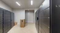 Located at 8800 Pine Forest Rd in Pensacola, Florida, this package and mail management room serves as a dedicated parcel receiving facility. The space is finished with large-format light beige tile flooring and bright white walls, creating a clean, functional environment illuminated by recessed and linear LED ceiling fixtures.

The room's most prominent feature is an extensive array of smart package lockers lining the walls on multiple sides. These dark matte metal units span floor to ceiling and are organized into numbered compartments of varying sizes, accommodating packages of different dimensions. A dedicated storage locker door is also integrated into the locker bank, providing secured access to additional storage.

A full-size commercial stainless steel refrigerator is positioned near the entry, likely used for the secure receipt of refrigerated or perishable deliveries — a notable and practical amenity. A community bulletin board is mounted nearby, displaying informational notices.

The room is accessible via a wood-toned door that connects to adjacent areas of the building, and a secondary doorway leads further into the facility. A closet or utility alcove with a hanging rod is also visible, adding additional functional storage to the space. Security cameras are installed, ensuring a monitored and safe environment for package retrieval. Overall, this room is a well-organized, modern package management solution designed for efficiency and resident convenience.