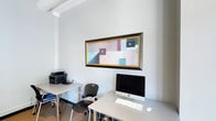 This single-floor workspace is designed with functionality and a clean aesthetic in mind. The interior is characterized by crisp white walls that create a bright, open atmosphere, complemented by an exposed beam detail near the ceiling that adds a subtle architectural element. The space is organized around practical workstations, including light-colored desks paired with an ergonomic mesh chair, providing a comfortable and efficient setup for daily tasks. A desktop computer workstation anchors one desk, while an adjacent surface accommodates a multifunction printer, making the layout well-suited for a productive work environment. The flooring features a neutral-toned carpet that adds warmth and reduces ambient noise. A standout decorative element is a large, horizontally oriented abstract painting mounted prominently on the wall, framed in black and gold. Its geometric composition in muted pastels, teal, and dark accents introduces a thoughtful artistic touch that balances the otherwise utilitarian setting. The overall ambiance is calm, orderly, and professional, making this space well-suited for focused individual work or a small team environment.