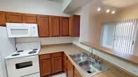 This single-floor residence is thoughtfully designed for everyday comfort and functionality. The kitchen serves as a central highlight of the space, showcasing warm-toned wood cabinetry that provides generous storage both above and below the countertops. The layout follows an efficient L-shape configuration, making the most of the available space while allowing for easy movement during meal preparation. A double-basin stainless steel sink is positioned conveniently along the counter, accompanied by a standard faucet. The kitchen is equipped with a white electric range and a built-in microwave, offering all the essentials for home cooking. Light-toned countertops complement the cabinetry, while tile flooring runs throughout, adding durability and ease of maintenance. A pass-through counter or breakfast bar area connects the kitchen to an adjacent space, enhancing the open feel of the layout. Track lighting on the ceiling ensures the workspace is well-lit, and a window with vertical blinds allows for natural light to filter into the area. The overall ambiance is clean, practical, and move-in ready, suited for those seeking a straightforward and well-equipped living space.