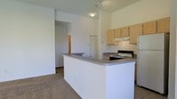 This single-floor residential space is designed with an open-concept layout that seamlessly connects the kitchen and living areas. The kitchen is equipped with light wood cabinetry, a built-in range with an overhead exhaust hood, and a spacious peninsula counter featuring a gray laminate surface — ideal for both meal preparation and casual dining. The peninsula effectively divides the kitchen from the adjoining living space, creating a natural flow between the two areas. The interior is finished with clean white walls and tall ceilings that contribute to an airy, open atmosphere. The living area is carpeted, while the kitchen transitions to tile flooring, providing a practical distinction between the two zones. A hallway visible from the main living area leads further into the unit, where a bi-fold door suggests a closet or utility space nearby. Track lighting and flush-mount ceiling fixtures provide ample illumination throughout. The overall design is straightforward and functional, offering a practical living environment with a neutral color palette that provides a versatile backdrop for personalization.