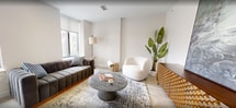 Located at 37 W Broad St in downtown Rochester, NY, this 710-square-foot single-floor apartment offers a thoughtfully designed open-concept layout combining the kitchen and living room into one cohesive space. The kitchen is outfitted with stainless steel appliances, including a full-size refrigerator and range with a sleek overhead hood vent, complemented by gray wood-tone cabinetry, white quartz countertops, and a classic subway tile backsplash. A peninsula with a white countertop and bar seating provides additional dining space and seamlessly connects the kitchen to the living area. Large windows throughout the unit invite abundant natural light and offer views of the surrounding Rochester cityscape. The living room features light wood-tone flooring, high ceilings, and a bright, neutral palette that creates an airy, open ambiance. The bathroom is finished with large-format tile flooring and a generously sized walk-in shower enclosed by sliding glass doors and lined with white subway tile, including a built-in bench. Throughout the unit, the clean architectural lines, light tones, and modern material choices contribute to a polished and contemporary urban living experience.