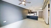 This single-floor space showcases a modern, open-concept design that seamlessly connects the living area and kitchen. The living space is defined by a bold, dark gray accent wall that creates a striking contrast against the lighter ceiling and flooring. A ceiling fan with a built-in light fixture is centrally positioned, providing both air circulation and ambient lighting throughout the space.

The kitchen is a standout feature, designed with dark cabinetry that extends from base to upper units, complemented by a light-colored countertop that wraps around to form a peninsula. This peninsula serves as both a workspace and a casual dining or gathering area. Stainless steel appliances, including a refrigerator and microwave, contribute to the kitchen's sleek, contemporary aesthetic. Pendant lighting hangs above the peninsula, adding focused illumination and a refined visual detail.

The flooring throughout is a neutral, light-toned surface that enhances the sense of openness. A secondary door is visible in the background, hinting at additional rooms or access points. Overall, the space conveys a clean, minimalist ambiance with a cohesive dark-and-light color palette.