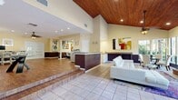This single-floor space offers a generous, open layout thoughtfully divided into distinct functional zones. The most striking architectural feature is the dramatic vaulted ceiling clad in warm wood planking, which crowns the living area and lends considerable vertical drama to the space. Recessed lighting throughout supplements the abundant natural light that pours in through large windows and glass-paneled French doors, offering views of lush outdoor greenery.

The living area is anchored by a full-height brick fireplace with a mounted television, surrounded by white upholstered seating and a glass-top coffee table atop a patterned area rug. Tile flooring defines this lounge zone, while a raised platform with wood-look flooring transitions seamlessly into a more active area featuring a glass-top desk and workstations along the perimeter—ideal for a business center or collaborative workspace.

A prominent counter with a dark paneled base and granite-style top serves as a reception or service area, dividing the entrance from the main gathering space. The hallway connects to additional back rooms, including what appears to be a fitness area with exercise equipment. Colorful abstract artwork displayed throughout adds a vibrant, contemporary aesthetic to the warm, neutral-toned interior.
