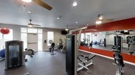 This fitness center is a purpose-built exercise facility designed to accommodate a wide range of workout routines. The space is outfitted with a variety of True-brand cardio machines, including an elliptical trainer and a stationary bike, providing options for low-impact cardiovascular exercise. A multi-functional strength training cable machine serves as a centerpiece for resistance workouts, complemented by free weights and additional exercise equipment visible throughout the room. One full wall is lined with large mirrors, accented by a bold red stripe, creating a sense of expanded space while allowing users to monitor their form during exercise. The flooring features durable rubber matting suited for high-traffic fitness use. Recessed lighting and multiple ceiling fans ensure the space remains bright and well-ventilated. Natural light filters in through windows fitted with blinds, adding to the open, energetic atmosphere. A wall-mounted television provides entertainment during workouts. The overall design is clean and functional, with a motivating, gym-like ambiance that supports both cardio and strength training goals.