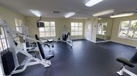 This dedicated fitness room offers a practical and well-organized space for strength training and exercise. The room is finished with durable rubber flooring, providing a safe and resilient surface suited for gym equipment and high-impact activity. Pale yellow walls create a bright, energizing atmosphere, complemented by abundant natural light streaming in through multiple double-pane windows that offer views of surrounding greenery.

The space is equipped with a selection of strength-training machines, including cable and resistance-based units, providing users with a range of workout options. A wall-mounted television is positioned for convenient viewing during exercise sessions. A large mirror spans one wall, enhancing the sense of space and allowing users to monitor their form while training.

Overhead fluorescent lighting ensures the room remains well-lit at all times, supplementing the natural light. A single exterior door provides direct access to the outdoors. The overall layout is open and uncluttered, allowing for ease of movement between equipment. The combination of functional amenities, natural light, and a clean, organized design makes this fitness room a practical and inviting space for daily workouts.