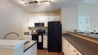 This 2-bedroom, 1-bathroom single-story residence offers a thoughtfully designed layout that balances functionality and comfort. The kitchen serves as a central highlight, featuring crisp white cabinetry with ample storage, dark countertops, and a full suite of black appliances including an electric range and a full-size refrigerator. A built-in range hood and track lighting overhead provide both practicality and a clean aesthetic. The kitchen flows seamlessly into the living area via a peninsula-style breakfast bar with seating, making it ideal for casual dining or entertaining. Wood-look flooring in the kitchen and dining transition gracefully into carpeted living and bedroom spaces, creating a warm, cohesive feel throughout. The living area is open and airy, benefiting from natural light and a neutral color palette that makes the space feel inviting. Both bedrooms are generously sized, each featuring plush carpeting, neutral walls, and closets with louvered doors. Large windows allow natural light to fill these rooms. The bathroom is well-finished with a white vanity, dark countertop, a large mirror, tile flooring in an earthy tone, and a lighted vanity bar. Clean white trim and neutral wall tones throughout the unit create a cohesive and move-in-ready environment.
