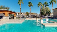 Located at 7606 W Peoria Ave in Peoria, Arizona, this vibrant outdoor amenity area showcases a resort-style design that captures the essence of Arizona living. The centerpiece is a generously sized, crystal-clear swimming pool with striking built-in water fountain features that add both visual appeal and a soothing ambiance. The pool deck is finished in textured concrete, providing ample space for lounging with multiple blue chaise lounge chairs and umbrella-shaded seating areas positioned throughout. A standout architectural feature is the covered ramada, built with stone-accented pillars and a tiled hip roof in the community's signature bold orange and neutral color palette, offering a shaded gathering space with additional seating. The surrounding landscape is lush with towering mature palm trees that frame the area and provide natural shade, complemented by desert-adapted shrubs and decorative rock, reflecting a classic Southwestern aesthetic. Iron fencing encloses the pool area for security, while the broader community buildings — featuring warm orange and tan exteriors with bold window trim accents — are visible in the background. The space is bathed in abundant Arizona sunshine, creating a bright, welcoming atmosphere ideal for outdoor relaxation and recreation year-round.