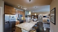 This single-floor residence offers two bedrooms and two bathrooms within a thoughtfully designed open-concept floor plan. The kitchen serves as a central focal point, showcasing granite countertops atop a generous island with bar seating, rich medium-toned wood cabinetry, a decorative tile backsplash, and stainless steel appliances including a refrigerator, range, and built-in microwave. Pendant lighting hangs above the island, complemented by a track lighting system that illuminates the workspace effectively.

The kitchen flows seamlessly into the dining and living areas, creating a cohesive entertaining space. Warm, neutral wall tones contrast with dark hardwood-style flooring, lending a grounded, modern aesthetic throughout the common areas. An arched architectural detail frames the transition into the living space, adding a classic design element. Decorative wall art and carefully placed lighting fixtures contribute to the polished, inviting ambiance. The open layout maximizes the sense of space, making the residence feel airy and connected throughout.