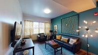 This single-floor residence combines contemporary design with vibrant personality, creating a cohesive and inviting living environment. The living room serves as the focal point of the space, anchored by a striking teal accent wall adorned with an oversized geometric gold wall sculpture. Dark hardwood-style flooring, a teal patterned area rug, and rich leather seating come together beneath natural light streaming through large windows dressed with bold golden-yellow patterned curtains. Curated wall art, including abstract canvases in blue and gold tones, adds an artistic dimension throughout the unit. The kitchen is functional and well-equipped, featuring warm honey-toned cabinetry, black appliances including a refrigerator, range, and microwave, and a sink with ample counter space. A compact dining table with modern chairs sits adjacent to the kitchen, offering a convenient dining area. The bedroom continues the teal accent wall theme, paired with plush carpeting, a lofted bed frame, a dedicated desk area, and a window that brings in natural light. The bathroom includes a wide dual-sink vanity with a large mirror, dark hardwood-style flooring, and a separate water closet, offering both practicality and comfort throughout.