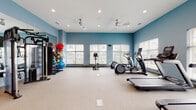 Located at 13915 S Autumn Way in Plainfield, IL, this fitness center offers a comprehensive, single-floor layout designed to support a full range of workout routines. The space is finished with calming blue-toned walls and neutral carpeted flooring, creating an inviting and energizing environment throughout.

The cardio zone is well-appointed with multiple treadmills, elliptical trainers, and a stationary bike, all arranged to maximize open floor space. Large windows with horizontal blinds line the walls, allowing generous natural light to fill the room while providing views of the surrounding exterior. Ceiling fans are installed throughout to maintain airflow and comfort during workouts.

The strength training area features a multi-functional cable machine by TAG, a full dumbbell rack with a wide range of weights, and a dedicated workout bench. A large wall mirror in this section enhances the sense of space and allows for proper form checks during exercise. A colorful medicine ball and stability ball rack adds versatility for functional fitness training.

Additional amenities include a mounted flat-screen television, a built-in water fountain station set against a decorative tiled accent wall, and a hands-free water bottle refill station. The space is thoughtfully designed to accommodate a variety of fitness levels and training styles in a clean, well-maintained facility.