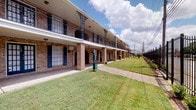 This property presents a well-maintained residential complex with a striking two-story brick exterior, accented by decorative ironwork, covered walkways, and a neatly landscaped lawn that runs along the length of the building. The exterior exudes a classic, functional character with arched brick columns and upper-level balconies accessible by a staircase at the end of the corridor.

The interior office space serves as the front-facing leasing or management area for the complex. Upon entry through elegant double glass French doors, visitors are greeted by a bright, open reception area featuring light wood-style flooring and a neutral gray color palette that lends a clean, professional atmosphere. The soaring ceiling height amplifies the sense of openness, and large windows allow abundant natural light to fill the space.

A comfortable seating area near the entrance, complete with an upholstered accent chair, provides a welcoming spot for visitors. Further inside, a dedicated workstation area with a dark wood desk and filing units serves as the administrative hub, neatly appointed for daily operations. Decorative framed artwork adorns the walls, adding a thoughtful finishing touch. The layout flows intuitively, balancing functional workspace with an inviting environment suited to greeting and assisting residents and prospective tenants.