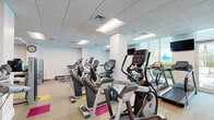 Located at 2500 Edwards Dr in Fort Myers, Florida, this well-equipped fitness center offers a generous, open exercise space designed to support a wide range of workout routines. The single-floor layout is brightened by recessed panel lighting set within a drop-tile ceiling, while large windows and glass doors along one wall invite natural light and provide views of the surrounding greenery outdoors.

The flooring features a neutral carpet base accented by vibrant pink and yellow tile insets, adding a lively, energetic character to the space. Cardio equipment is well-represented, with multiple Matrix-brand treadmills, elliptical cross-trainers, and stationary bikes arranged in orderly rows, each unit equipped with individual monitors for entertainment during workouts.

Strength training is thoroughly addressed through a dedicated area housing a full dumbbell rack, adjustable weight benches, cable pull stations, and a variety of selectorized resistance machines targeting multiple muscle groups. A rack of stability and medicine balls of varying sizes rounds out the functional fitness offerings.

Wall-mounted televisions provide entertainment throughout the space, and a wall clock keeps users on schedule. White-painted structural columns are integrated throughout the open floor plan, maintaining a clean, modern aesthetic. The overall ambiance is bright, organized, and purpose-built for effective fitness training.
