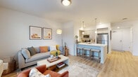 This single-floor residence offers a thoughtfully designed open-concept layout that seamlessly connects the living, dining, and kitchen areas. Light-toned hardwood-style flooring flows throughout the main living spaces, complementing the crisp white walls and creating a clean, contemporary aesthetic.

The kitchen is well-appointed with white shaker-style cabinetry, quartz countertops, and a full suite of stainless steel appliances, including a refrigerator, range, microwave, and dishwasher. Pendant lighting hangs above the kitchen island, which features bar seating and serves as a natural divider between the kitchen and living area.

The living room benefits from generous natural light through large windows and offers comfortable space for both lounging and entertaining. A dedicated hallway connects the main living area to the private bedroom, which features plush carpeting, additional windows with outdoor views, and ample room for furnishings.

A dedicated laundry room with a full-size side-by-side washer and dryer adds everyday convenience, along with wire shelving for storage. Throughout the unit, the design emphasizes clean lines, neutral tones, and functional use of space, resulting in a bright and welcoming atmosphere.