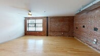 Located at 1700 Fairview St in Greensboro, NC, this 2-bedroom, 1-bathroom loft-style residence showcases a compelling blend of industrial character and modern finishes across a single floor. The open living room is anchored by striking exposed brick walls, light hardwood flooring, and a ceiling fan, with windows allowing natural light to brighten the space. The kitchen is well-equipped, featuring rich dark-espresso cabinetry, granite-look countertops, a double stainless steel sink, and a full suite of black appliances including a refrigerator, range, microwave, and dishwasher. Exposed ductwork overhead adds to the loft's industrial aesthetic. The primary bedroom continues the exposed brick motif, complemented by plush carpeting, a ceiling fan, and ample room to accommodate furnishings. The second bedroom also features carpet flooring and a ceiling fan, offering a comfortable and versatile space. Three hallways connect the layout efficiently, while two closets provide dedicated storage. The bathroom is clean and functional, finished with marble-look tile flooring, a soaking tub with shower, and chrome fixtures. The overall ambiance is distinctly urban, combining raw architectural elements with thoughtful modern updates throughout.