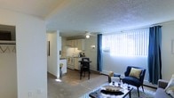 This single-floor residence offers a functional and open layout that integrates the living, dining, and kitchen areas into one cohesive space. The living area is carpeted and comfortably accommodates a seating arrangement, anchored by a glass-top coffee table. Large windows dressed with blue curtains allow generous natural light to fill the room, creating a bright and welcoming ambiance. A ceiling fan overhead contributes to year-round comfort. The dining area sits at the heart of the space, furnished with a compact bistro-style table and chairs, making it ideal for casual meals. The kitchen flows directly from the dining area and features white cabinetry, a white refrigerator, and a matching white range with overhead ventilation — all laid out for efficient use. The kitchen also benefits from its own ceiling fan and connects to an additional area visible through a doorway, suggesting extra storage or a utility space. The textured ceiling is consistent throughout, adding a subtle design element to the interior. The overall aesthetic is clean and neutral, with light-toned walls that provide a versatile backdrop. This well-connected, single-level layout is practical and easy to navigate.
