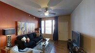 This well-appointed single-floor residence showcases a thoughtfully designed living area that blends comfort with contemporary style. The living room serves as the centerpiece of the space, featuring rich hardwood-style flooring that anchors the room with warmth and elegance. A bold terracotta accent wall adds a striking visual contrast, complemented by a large abstract canvas artwork that enhances the room's artistic character. Large windows dressed with dark curtains allow generous natural light to fill the space, creating a bright and welcoming atmosphere. A ceiling fan with a built-in light fixture provides both functionality and a classic finishing touch overhead. The living area is furnished with a plush dark sofa accented with decorative throw pillows, paired with a sleek glass-and-gold coffee table atop a patterned area rug, offering a modern yet cozy seating arrangement. A flat-screen television setup completes the entertainment area. The entry door is conveniently situated within the living space, offering ease of access. Overall, the space presents a cohesive and inviting interior design with warm tones, practical finishes, and a layout well-suited for everyday living.