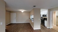 This single-floor residence offers a clean, functional layout centered around an open-concept living and kitchen area. The living space is finished with warm beige walls, a textured ceiling, and rich hardwood-style flooring that extends throughout, creating a cohesive and inviting atmosphere. A ceiling fan with light fixture provides both comfort and illumination, while a standard entry door with a classic white panel design offers a welcoming entrance.

The kitchen is separated from the living area by a half-wall partition featuring a dark countertop breakfast bar, providing a natural division between spaces while maintaining an open feel. The kitchen is well-equipped with stainless steel appliances, including a gas range and over-the-range microwave, complemented by crisp white cabinetry that maximizes storage. The cabinetry extends to the upper walls, offering ample space for organization.

Neutral-toned walls throughout the space create a versatile backdrop suitable for a variety of interior design styles. HVAC vents are positioned in the ceiling to ensure efficient climate control. Overall, the space strikes a balance between practicality and comfort, making it a straightforward yet functional single-floor living environment.