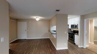 This single-floor residence offers a clean, functional layout centered around an open-concept living and kitchen area. The living space is finished with warm beige walls, a textured ceiling, and rich hardwood-style flooring that extends throughout, creating a cohesive and inviting atmosphere. A ceiling fan with light fixture provides both comfort and illumination, while a standard entry door with a classic white panel design offers a welcoming entrance.

The kitchen is separated from the living area by a half-wall partition featuring a dark countertop breakfast bar, providing a natural division between spaces while maintaining an open feel. The kitchen is well-equipped with stainless steel appliances, including a gas range and over-the-range microwave, complemented by crisp white cabinetry that maximizes storage. The cabinetry extends to the upper walls, offering ample space for organization.

Neutral-toned walls throughout the space create a versatile backdrop suitable for a variety of interior design styles. HVAC vents are positioned in the ceiling to ensure efficient climate control. Overall, the space strikes a balance between practicality and comfort, making it a straightforward yet functional single-floor living environment.
