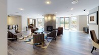 This thoughtfully designed single-floor amenity space offers a welcoming and functional environment across several distinct zones. The main lounge area serves as the heart of the space, featuring rich hardwood-style flooring, recessed and track lighting, and a striking floor-to-ceiling tiled fireplace as a central focal point. Comfortable seating arrangements, accented by vibrant chevron-patterned area rugs, create an inviting atmosphere for relaxation or socializing. Expansive floor-to-ceiling glass doors and windows flood the lounge with natural light and provide views of an outdoor patio area, seamlessly connecting the interior to the exterior. A secondary lounge zone offers additional seating with upholstered armchairs and a round coffee table, ideal for smaller gatherings. The kitchen is fully equipped with stainless steel appliances, including a gas range, over-the-range microwave, and refrigerator, complemented by light wood cabinetry and a practical kitchen bar with seating. A billiards table adds a recreational element to the space. Glass-paneled interior doors provide visual connectivity between areas while maintaining defined spaces. Overall, the space balances modern design with warm, functional details suited for a community or shared-use setting.