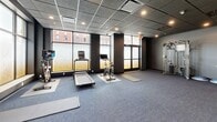 Located at 224 N Barstow St in Eau Claire, Wisconsin, this fitness center offers a thoughtfully designed, single-floor layout dedicated to health and wellness. The spacious exercise room is the heart of the facility, featuring a dark, drop-panel ceiling with recessed lighting that creates a focused and energizing atmosphere. Dark gray walls and durable carpet tile flooring lend the space a sleek, contemporary aesthetic. A structural column with a distinctive textured finish serves as an architectural focal point within the open floor plan. Large, frosted-panel windows line the walls, allowing natural light to filter in while maintaining privacy, and offering partial views of the surrounding Eau Claire streetscape. The exercise room is equipped with treadmills, stationary cycling bikes, a cable machine, free weights, benches, and exercise mats, providing a comprehensive range of workout options. A wall-mounted mirror on one end enhances the sense of space. The facility also includes a water fountain near the entrance for added convenience. The adjacent bathroom is clean and modern, finished with white subway tile wainscoting, large-format floor tiles, and black matte hardware accents. It is fully ADA-accessible, featuring grab bars and a built-in bench. The overall design of the facility is functional, modern, and well-suited for regular fitness use.