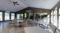 This single-floor gathering space is designed to accommodate communal dining and socializing in a comfortable, well-appointed setting. The room features a striking vaulted ceiling with a crisp white finish, anchored by a ceiling fan and complemented by recessed lighting that ensures the space is well-lit at any hour. Contrasting dark-toned walls lend a warm, grounded ambiance that balances the brightness of the ceiling above.

Large sliding glass doors and expansive windows line the perimeter of the room, flooding the interior with natural light and offering unobstructed views of the lush, tree-lined outdoor surroundings. These generous openings also provide a seamless visual connection between the interior and the exterior, creating an open, airy atmosphere.

The dining area is furnished with multiple tables and lattice-back chairs in a neutral palette, arranged to accommodate both small groups and larger gatherings with ease. The light-toned flooring further enhances the sense of openness throughout the space. Decorative wall art adds a subtle touch of personality to the room. Emergency exit signage and thoughtful layout details reflect attention to both safety and functionality, making this a practical yet inviting space for everyday use.