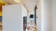 This mail and package room is a thoughtfully designed communal space that balances practicality with modern aesthetics. The room is defined by its striking geometric-patterned tile flooring, featuring an eye-catching arrangement of hexagonal and angular shapes in tones of white, grey, and brown that runs throughout the entire space, creating a bold visual foundation.

Rows of matte black individual mailboxes line the walls, extending the full length of the room and providing ample mail storage. A dedicated package area features additional black lockers at the far end, set against a decorative accent wall adorned with an intricate geometric wallpaper pattern in black and white, adding a layer of visual interest to the functional space.

Overhead, a series of brass pendant lights hang at varying heights, casting warm, focused illumination down the corridor-style layout. Recessed ceiling lights supplement the pendant fixtures, ensuring the space is well-lit throughout.

Floor-to-ceiling black-framed glass windows and a matching glass entry door flood the room with natural light and offer views of the surrounding greenery and outdoor areas. The crisp white walls and clean architectural lines complete the contemporary design, making this a polished and welcoming utility space.