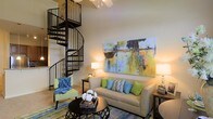 This two-floor space makes an immediate impression with its dramatic spiral staircase, a standout architectural feature crafted in sleek black metal that winds elegantly upward and serves as both a functional element and a visual centerpiece. The living area benefits from soaring ceilings that amplify the sense of openness and volume throughout the space. A ceiling fan is positioned overhead, adding both comfort and a casual, airy feel to the room. The living area is warmly lit with ambient lighting that complements the neutral wall tones, creating an inviting and relaxed atmosphere. Large-scale abstract wall art in vivid blues, greens, and yellows adds a bold, contemporary focal point to the main living space. The flooring features plush carpeting accented by a patterned area rug in cool blue and green tones, tying together the overall color palette. The layout accommodates comfortable seating and allows for easy flow between areas. The overall design balances modern accents with warm, approachable furnishings, making the space feel both stylish and livable. The second floor, accessible via the spiral staircase, adds functional versatility and contributes to the home's distinctive, multi-dimensional character.