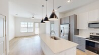 This well-appointed single-story residence in Austin, TX offers a thoughtfully designed open-concept layout that seamlessly connects the kitchen, dining, and living areas. Light hardwood flooring flows throughout the main living spaces, complemented by an abundance of natural light streaming through multiple windows and a glass exterior door.

The kitchen is a standout feature, centered around a large island with a quartz countertop and undermount sink, illuminated by a trio of modern black pendant lights. Two-tone cabinetry — white uppers paired with gray lowers — lines the walls, accompanied by a classic subway tile backsplash and a full suite of stainless steel appliances, including a French door refrigerator, range, and microwave.

The living area is bright and open, featuring a ceiling fan and access to the outdoors via a glass-paneled door. The bedroom is carpeted and well-lit by a window with natural light, offering direct access to both the walk-in closet and the bathroom. The walk-in closet provides built-in shelving and hanging rods for organized storage.

The bathroom features gray cabinetry with quartz counters, marble-style tile flooring, a soaking tub, and a separate glass-enclosed shower, creating a clean and functional space.