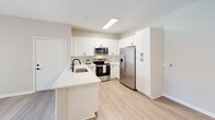 This thoughtfully designed one-bedroom, one-bathroom unit in Murrieta, California offers a clean, modern aesthetic with functional living spaces throughout. The kitchen is a standout feature, showcasing crisp white shaker cabinetry that extends to the ceiling for ample storage, a hexagonal tile backsplash, and a suite of stainless steel appliances including a French door refrigerator, electric range, and over-the-range microwave. A peninsula with a white quartz countertop and a matte black undermount sink adds both workspace and a casual dining option, while light wood-tone flooring ties the area together seamlessly. The bedroom offers a comfortable and tranquil retreat, complete with plush carpeting, a ceiling fan, and a window that frames views of an outdoor pool area, allowing natural light to fill the room. A dedicated closet provides practical storage. The bathroom is finished with a white vanity, a large mirror with contemporary lighting, matte black hardware accents, and wood-look flooring, paired with a full shower-tub combination. A hallway connects the spaces efficiently, contributing to the unit's smart, compact layout. Neutral wall tones and consistent modern finishes create a cohesive, inviting atmosphere throughout.