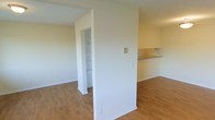 This single-floor residence offers a clean, straightforward layout designed for comfortable everyday living. The interior is finished with warm hardwood-style flooring that extends throughout the open living area, lending a cohesive and inviting feel to the space. Neutral, light-toned walls reflect natural light effectively, contributing to a bright and airy atmosphere throughout.

A defining architectural feature is the partial dividing wall that separates the living area from the kitchen zone, where a countertop pass-through provides a convenient connection between the two spaces. This design element allows for an open feel while still maintaining a sense of defined areas for living and food preparation.

Towards the back of the living area, a dedicated built-in storage nook—finished with white shelving—offers practical organizational space, ideal for household essentials or linens. White baseboards and trim throughout add a crisp contrast to the warm flooring beneath.

The overall ambiance of the space is clean, minimal, and move-in ready, offering a functional foundation that is easy to personalize. The straightforward design and well-maintained finishes make this an accessible and practical space suited to a range of lifestyles.