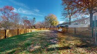 Located at 10035 Dabney Dr in Charlotte, North Carolina, this outdoor dog park offers a generously sized, ground-level green space designed for pets and their owners. The area is fully enclosed by a modern horizontal-slat wood fence, constructed with natural cedar planks mounted on sleek black steel posts. This contemporary fencing design provides both a secure boundary and an aesthetically appealing perimeter that complements the natural surroundings.

The grounds are predominantly grassy, offering ample room for pets to roam and play freely. Mature deciduous trees are scattered throughout and along the perimeter, providing natural shade and seasonal color, as evidenced by the warm autumn foliage visible throughout the space. White Adirondack chairs are positioned within the enclosure, giving pet owners a comfortable place to relax while supervising their animals.

A dedicated waste station with signage and a refuse container is conveniently situated within the park, promoting cleanliness and responsible pet ownership. The open layout benefits from abundant natural light and wide views of the sky, creating a bright and welcoming atmosphere. The surrounding neighborhood is visible beyond the fencing, lending a sense of community to this well-maintained outdoor amenity.