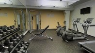 This fitness center is a dedicated exercise space designed to support a well-rounded workout routine. The room features rubberized flooring, which provides both safety and comfort underfoot, and is finished with warm yellow-toned walls that contribute to an energizing atmosphere. Overhead fluorescent lighting ensures the space is brightly and evenly illuminated throughout.

The strength training area is anchored by a cable machine/functional trainer, ideal for a variety of resistance exercises, accompanied by an adjustable weight bench and a set of dumbbells arranged along one wall. Full-length mirrors line the adjacent wall, allowing users to monitor their form during exercises and giving the space a more open, expansive feel.

The cardio section includes a Life Fitness elliptical cross-trainer and a stationary upright bike, offering effective low-impact cardiovascular workout options. Informational notices are posted on the wall, likely detailing facility rules and equipment guidelines for users.

Overall, this fitness center is thoughtfully organized to maximize the use of its footprint, offering a functional and practical environment for both strength and cardiovascular training. The layout allows for comfortable movement between equipment, making it a convenient and accessible amenity for regular use.